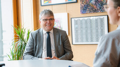 Ein Mann im Anzug sitzt im Büro und lächelt eine Interviewerin an, die ihm gegenübersitzt