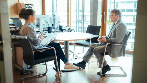 Ein Mann im Anzug in einem großen verglasten Raum sitzt an einem Tisch einer Interviewerin gegenüber